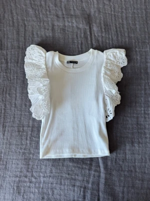 Vit topp med volangärmar från Zara - Supersöt vit topp från Zara med ribbat material och stora volangärmar i broderad spets. Perfekt fina sommar outfits! Toppen har en croppad passform och rund halsringning. Bra i storlek!