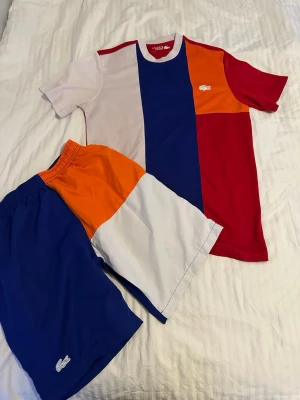 Färgglad pyjamas från Lacoste - Säljer ett set pyjamas från Lacoste med t-shirt och shorts i färgblocksmönster. T-shirten har rött, blått, vitt och orange medan shortsen matchar med samma färger. Båda plaggen har Lacoste-loggan och är tillverkade i mjukt bomullsmaterial.