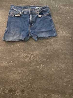 LOW Rise Blå jeansshorts från Levi's - Säljer ett par klassiska blå jeansshorts från Levi's med rå kant och fem fickor. Shortsen har normal passform och knappgylf. Perfekta för varma dagar och en avslappnad stil.