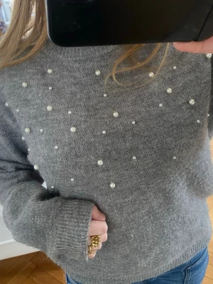 Grå stickad tröja med pärlor - Mysig grå stickad tröja dekorerad med små vita pärlor på framsidan. Tröjan har rund halsringning, långa ärmar och en avslappnad passform. Perfekt för dig som gillar en stilren men ändå trendig look.