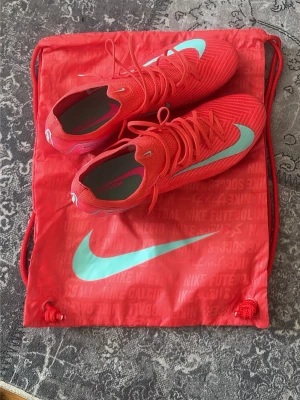 Nike Mercurial elite - Säljer ett par röda Nike Mercurial Elite FG med turkosblå swoosh. Skorna har snörning och är tillverkade i ett lätt syntetmaterial som ger bra bollkontroll och passform. Perfekta för dig som vill sticka ut på planen med både stil och funktion. Endast används under ett inhopp på 35 min.