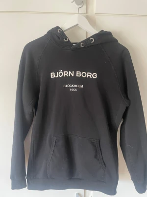 Svart hoodie från Björn Borg - Svart hoodie från Björn Borg med vit text på bröstet där det står 'BJÖRN BORG STOCKHOLM 1956'. Hoodien har huva med dragsko och en stor magficka. Mjuk insida och ribbade muddar vid ärmslut och nederkant. Perfekt för en chill och sportig stil.