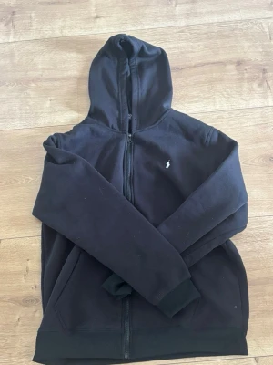 Svart hoodie från Ralph Lauren - Svart hoodie med dragkedja från Ralph Lauren. Klassisk design med huva, långa ärmar och vit broderad logga på bröstet. Tillverkad i mjukt bomullsmaterial som är skönt att ha på sig. Perfekt för en avslappnad och stilren look.