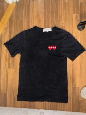 Svart Comme des Garçons Play t-shirt - Svart t-shirt från Comme des Garçons Play med två röda hjärtan med ögon broderade på bröstet. Klassisk rund hals och korta ärmar. Perfekt för dig som gillar streetwear och vill ha något stilrent.                                       Köpte originalt för runt 1200kr jag har använt ganska ofta sen dess allt funkar bra size M