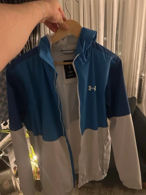 Blå och vit vindjacka från Under Armour - Snygg vindjacka från Under Armour i blått och vitt med dragkedja och huva. Jackan har meshfoder på insidan och logga på bröstet. Perfekt för blåsiga dagar och sportiga outfits.