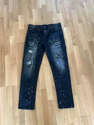 Svarta Dsquared2 jeans med slitningar - Säljer ett par svarta jeans från Dsquared2 med coola slitningar och färgstänk. Jeansen har raka ben, klassisk femficksmodell och en tydlig Dsquared2-tag vid gylfen. Materialet är denim med stretch för extra komfort.