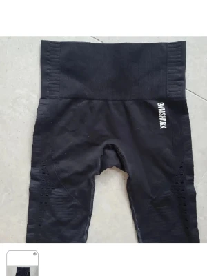 Svarta seamless tights Gymshark - Svarta seamless leggings från Gymshark med hög midja och ribbade detaljer längs sidorna. Loggan är tryckt i vitt på ena höften. Stretchigt material som sitter tight och formar sig efter kroppen. Perfekta för träning eller chill.
