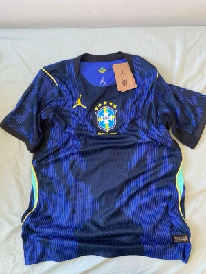 Blå Brasilien matchtröja Nike - Snygg blå matchtröja från Nike för Brasiliens landslag. Tröjan har gula detaljer, CBF-emblem på bröstet och Jordan-logga. Tillverkad i lätt och ventilerande funktionsmaterial, perfekt för fotboll eller träning.