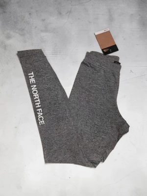 Grå leggings från The North Face - Snygga grå leggings från The North Face med vit logga längs ena benet. De har en smal passform och är tillverkade i ett mjukt och stretchigt material som sitter skönt hela dagen. Perfekta för en sportig och avslappnad stil.