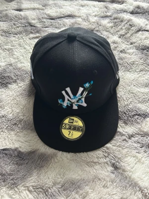 Svart New Era Yankees keps 59Fifty - Säljer en svart New Era 59Fifty keps med New York Yankees-logga framtill dekorerad med blå blommor. Kepsen har platt skärm och broderad detalj på sidan. Klassisk streetwear-look och riktigt snygg till vardagsoutfiten. Skick 10/10