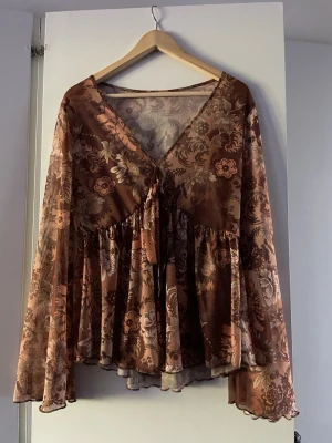 Brun blommig blus med vida ärmar - Snygg brun blus med blommigt mönster och v-ringning. Blusen har knytning framtill och vida, långa ärmar i ett lätt, slitsad vid magen och aldrig använd🌸😀