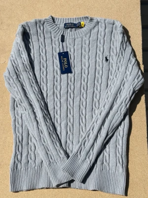 Ljusgrå kabelstickad tröja Polo Ralph Lauren - Ljusgrå kabelstickad tröja från Polo Ralph Lauren med rund halsringning och ribbade muddar. Klassisk broderad logga på bröstet och mjukt material som känns skönt mot huden. Perfekt för dig som gillar stilrena och tidlösa plagg.