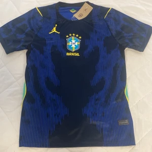 Blå Brasilien fotbollströja Nike - Snygg blå fotbollströja från Nike med Brasilien-logga och gula detaljer. Tröjan har korta ärmar, rund hals och ett unikt mönster i mörkblått. Perfekt för fotbollsträning eller att bära som supporter. Materialet är lätt och andas, vilket gör den bekväm under aktivitet.