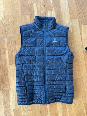 Mörkblå dunväst från Haglöfs - Snygg mörkblå dunväst från Haglöfs med quiltad design och hög krage. Västen har dragkedja framtill och diskret logga på bröstet. Perfekt för lager-på-lager och outdoor-looks. Lätt och smidig att bära med sig.