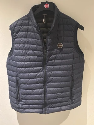 Marinblå dunväst från Colmar - Snygg marinblå dunväst från Colmar med quiltad design och dragkedja framtill. Storleken ligger mellan medium och small, på mig som personligen varierar mellan medium och large är västen lite liten. Västen har två praktiska fickor med dragkedja och en diskret logotyp på bröstet. Perfekt för lager-på-lager och stilren look. Västen har även en inneficka på vänster sida. Perfekt skick och sällan använd, inga defekter. Hör av dig vid minsta fundering!