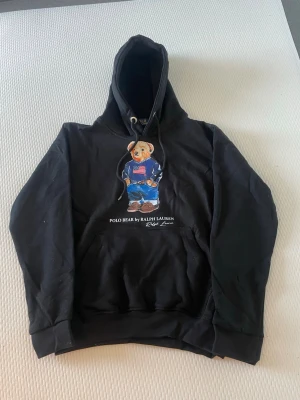 Svart hoodie Polo Ralph Lauren S - Svart hoodie från Polo Ralph Lauren med den ikoniska Polo Bear-trycket på bröstet. Hoodien har dragsko i huvan och en klassisk känguruficka framtill. Tillverkad i mjukt bomullsmaterial som är skönt att bära. Perfekt för en avslappnad och stilren look.