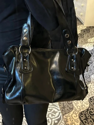 Svart handväska skinn - Snygg svart handväska i blankt skinnimitation med dubbla handtag och silverfärgade metalldetaljer. Väskan har en klassisk rektangulär form och dekorativa nitar på handtagen. 