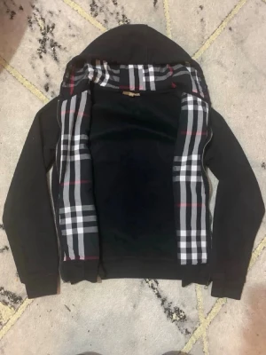 Svart Burberry hoodie med rutigt foder storlek M - Svart hoodie från Burberry med klassiskt rutigt foder i huvan och längs insidan. Dragkedja framtill och långa ärmar. Perfekt för dig som vill ha en stilren hoodie med en touch av lyx och ikoniskt mönster.
