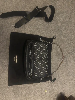 Svart handväska från Zadig & Voltaire - Cool svart handväska i skinn från Zadig & Voltaire med silverfärgade nitar i zigzag-mönster. Väskan har både en kedjehandtag och en avtagbar axelrem i textil, samt dragkedja framtill. Perfekt accessoar för att lyfta din outfit.