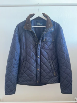 Mörkblå quiltad jacka från polo ralph lauren - Quiltad mörkblå jacka från polo ralph lauren med bruna detaljer på kragen och tryckknappar. Jackan har en bröstficka med knapp, två sidofickor och dragkedja framtill. Perfekt för kyligare dagar och har en klassisk, stilren look.