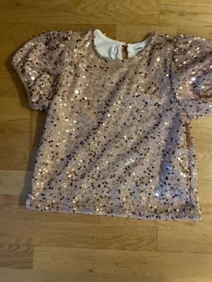 Glittrig paljettopp från Sinsay - Supersnygg topp från Sinsay täckt med skimrande paljetter i roséguld. Puffiga korta ärmar och rund halsringning. Perfekt för dig som vill sticka ut med extra glitter och glam. Materialet är mjukt på insidan för skön känsla mot huden.