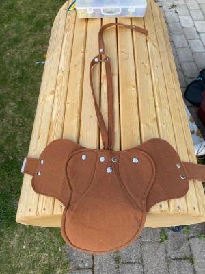 Brun tygsadel till leksakshäst  - Säljer en brun sadel i tyg med silverfärgade nitar, designad för käpphäst eller ponnyhäst. Sadelns form är anpassad för att sitta stabilt och har remmar för enkel fastsättning. Materialet är mjukt tyg och färgen är varmbrun. Perfekt för maskerad, cosplay eller hobby med käpphästar eller leksakshäst. 
