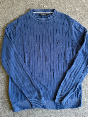 Blå stickad tröja från Nautica - Säljer en blå stickad tröja från Nautica med rund halsringning och diskret logga på bröstet. Tröjan har ett ribbat mönster längs hela plagget samt ribbade muddar vid ärmslut och nederkant. Perfekt för lager-på-lager och snygg till jeans.