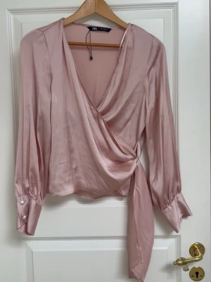 Rosa satin omlottblus - En ljusrosa omlottblus från Zara i satin material. Blusen har långa ärmar med breda manschetter och pärlknappar samt knytband i midjan. 