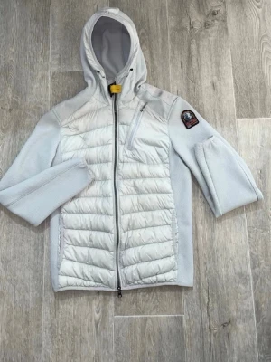 Vit pufferjacka från Parajumpers - Snygg vit pufferjacka från Parajumpers med huva och dragkedja framtill. Jackan har quiltade partier på framsidan, en bröstficka med dragkedja och klassisk PJS-logga på ärmen. Perfekt för kyliga dagar och har en sportig look.