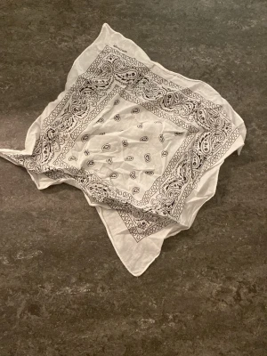Vit bandana med paisleymönster - Snygg vit bandana med klassiskt svart paisleymönster. Perfekt accessoar att knyta runt halsen eller i håret för en cool och avslappnad stil. Tunn och lätt i materialet, passar till många outfits.