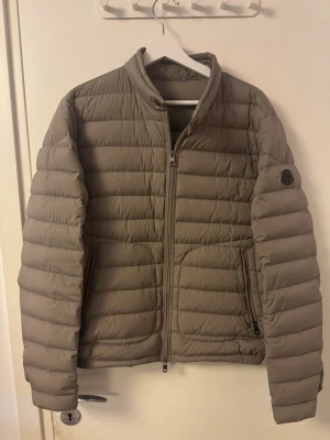 Beige dunjacka från Moncler - Snygg beige dunjacka från Moncler med klassisk quiltad design och dragkedja framtill. Jackan har två fickor med dragkedja och Moncler-logga på ärmen. Perfekt för kalla vinterdagar och riktigt stilren look.Moncler Acorus beige. Storlek: 3(M/L). Skick: 8/10.
