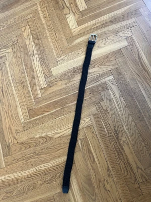 Svart flätat bälte med silverfärgat spänne 80cm - Snyggt svart flätat bälte med ett klassiskt silverfärgat spänne och detaljer i skinn. Perfekt accessoar för att lyfta din outfit och funkar till både jeans och kostymbyxor.
