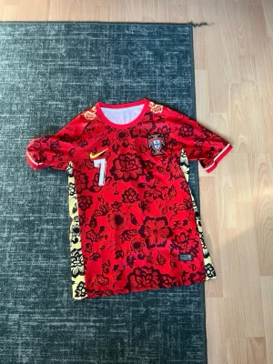 Portugal Ronaldo matchtröja röd - Säljer en röd Portugal matchtröja från Nike med blommigt mönster, gula detaljer på sidorna och nummer 7 samt Ronaldo på ryggen. Tröjan har korta ärmar och Portugal-emblemet på bröstet. Perfekt för dig som älskar fotboll och vill sticka ut på planen.