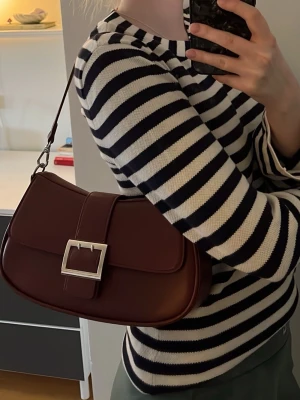 Vinröd axelväska med silverdetalj - Snygg vinröd handväska i slätt konstläder med axelrem och stor silverfärgad spänndetalj framtill. Väskan har en stilren, avlång form och minimalistisk design som passar perfekt till trendiga outfits. Använd fåtal gånger, mycket gott skick💕💌