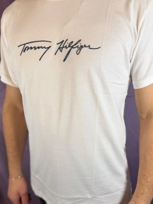 Vit T-shirt från Tommy Hilfiger - Snygg vit T-shirt från Tommy Hilfiger med svart logotyp i skrivstil på bröstet. Klassisk passform och rund halsringning. Tillverkad i mjuk bomull som känns skön mot huden. Perfekt för en clean och stilren look.