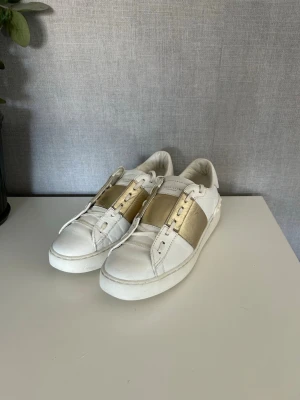 Valentino opens  - Snygga vita sneakers från Valentino med bred guldfärgad rem över vristen och vita snören. Skorna har en chunky sula och är tillverkade i läder. Ikoniska nitar baktill ger extra edge till looken. Perfekta för dig som vill sticka ut med stil.
