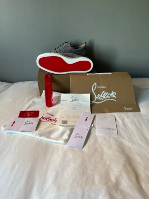 Louboutin skor - Snygga gråa sneakers från Christian Louboutin i mocka med klassisk röd sula och vita kanter. Skorna har lågt skaft, rund tå och matchande grå skosnören. Perfekta för dig som vill sticka ut med exklusiva detaljer och lyxig känsla.