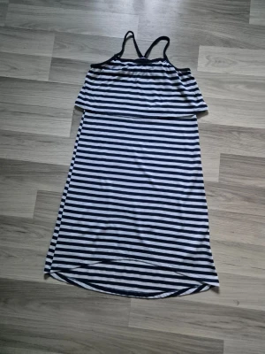 Randig sommarklänning med smala axelband - Snygg klänning med marinblå och vita horisontella ränder. Modellen har smala axelband och en lös, rak passform som slutar strax under knäet. Perfekt för varma dagar och enkel att styla. Materialet känns mjukt och lätt.