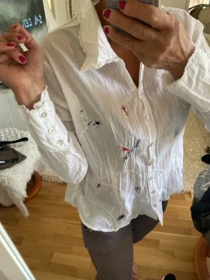 Vit skjorta med broderade fjärilar - Snygg vit skjorta med broderade fjärilar i olika färger på framsidan. Skjortan har lång ärm, klassisk krage och knäppning framtill. Materialet är tunt och lätt, perfekt för en avslappnad och trendig look.