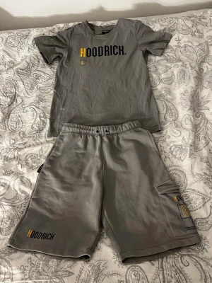 Grått Hoodrich set med t-shirt & shorts - Säljer ett grått set från Hoodrich med t-shirt och matchande shorts. T-shirten har tryckt logga i svart och gult både fram och bak, och shortsen har elastisk midja samt logga på benet och ficka. Perfekt för en avslappnad streetwear-look.