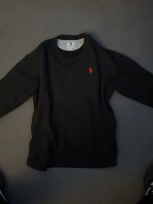 Svart stickad tröja U.S. Polo Assn. - Svart stickad tröja från U.S. Polo Assn. med röd broderad logga på bröstet. Tröjan har rund halsringning och långa ärmar. Perfekt för dig som gillar klassisk och stilren design.