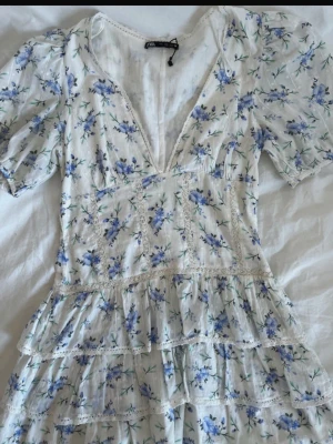 Blommig volangklänning från Zara - Romantisk vit klänning från Zara med blått blommönster, djup v-ringning och volangdetaljer. Klänningen har korta puffärmar och är sydd i ett lätt bomullstyg, perfekt för varma dagar. Söt spetskant längs volangerna ger extra charm.
