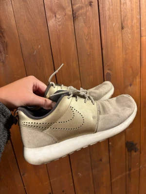 Beige Nike Roshe One sneakers - Snygga beige Nike Roshe One sneakers med vit sula och diskret perforerad swoosh på sidan. Skorna har lågt skaft, grå snörning och svart innerfoder. Perfekta för en avslappnad och stilren look.
