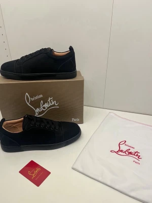 Svarta sneakers från Christian Louboutin - Stilrena svarta sneakers från Christian Louboutin i mocka med svarta snören och diskret logga. Låg modell med klassisk siluett och beige innersula. Perfekta för dig som gillar exklusiva och minimalistiska skor.
