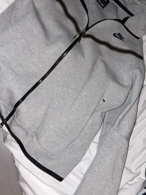 Grå zip hoodie från Nike - Snygg grå hoodie från Nike med dragkedja framtill och svart logga på bröstet. Hoodien har svarta och bruna detaljer längs bröstet och en klassisk huva. Perfekt för en sportig och avslappnad stil.