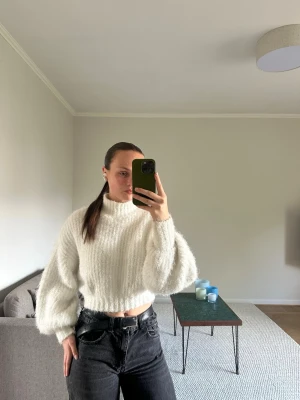 Vit croppad stickad tröja - Säljer en fluffig, vit croppad stickad tröja med ballongärmar och hög hals. Tröjan har en mjuk och mysig känsla, perfekt för kyligare dagar. Passar snyggt till både jeans och kjol för en trendig look.
