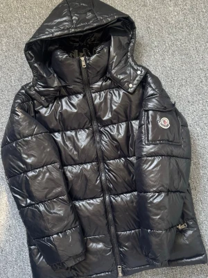Svart dunjacka från Moncler med huva - Moncler Maya jacka i svart, NFC märke tillgängligt. Jackan har Moncler-logga på ärmen och praktiska fickor. 