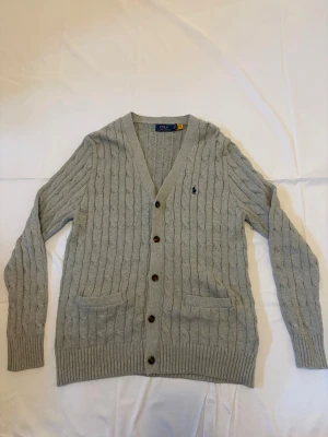 Grå kabelstickad kofta Polo Ralph Lauren - Grå kabelstickad cardigan från Polo Ralph Lauren. Tröjan är i mycket gott skick utan synliga defekter, fläckar eller slitage. Detaljer: 	•	Märke: Polo Ralph Lauren 	•	Modell: Cable Knit Cardigan 	•	Färg: Grå melange med marinblå inbrodererad pony-logga 	•	Storlek: S 	•	Passform: Regular 	•	V-ringning med bruna hornknappar 	•	Två fickor 	•	Material: Bomull Skick: Mycket gott — inga defekter.                     Modellen: 178 cm 
