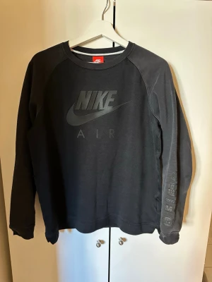 Svart Nike Air sweatshirt - Svart sweatshirt från Nike Air med stor logga framtill och tryck på ena ärmen. Tröjan har rund halsringning, raglanärmar och är gjord i mjukt bomullsmaterial. Perfekt för en sportig och avslappnad stil.