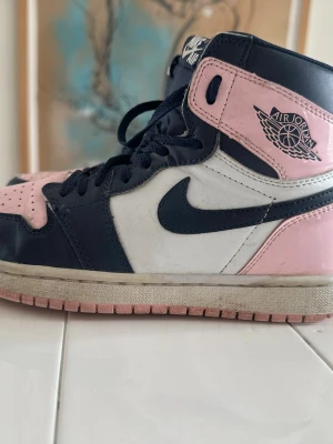 Air Jordan 1 Retro High OG Pink/Marin - Snygga Air Jordan 1 Retro High OG sneakers i rosa, marinblått och vitt läder. Klassisk hög siluett med svart swoosh och Air Jordan-logga på sidan. Perfekta för dig som vill sticka ut med en ikonisk och trendig sneaker.